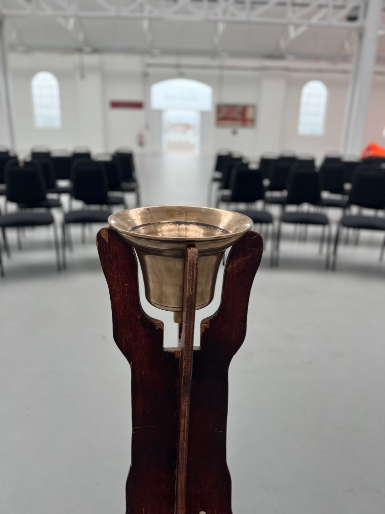 In foreground a brass ship's bell is held upturned in a wooden stand, to the rear black chairs are arranged in a horseshoe against the white background of the building..